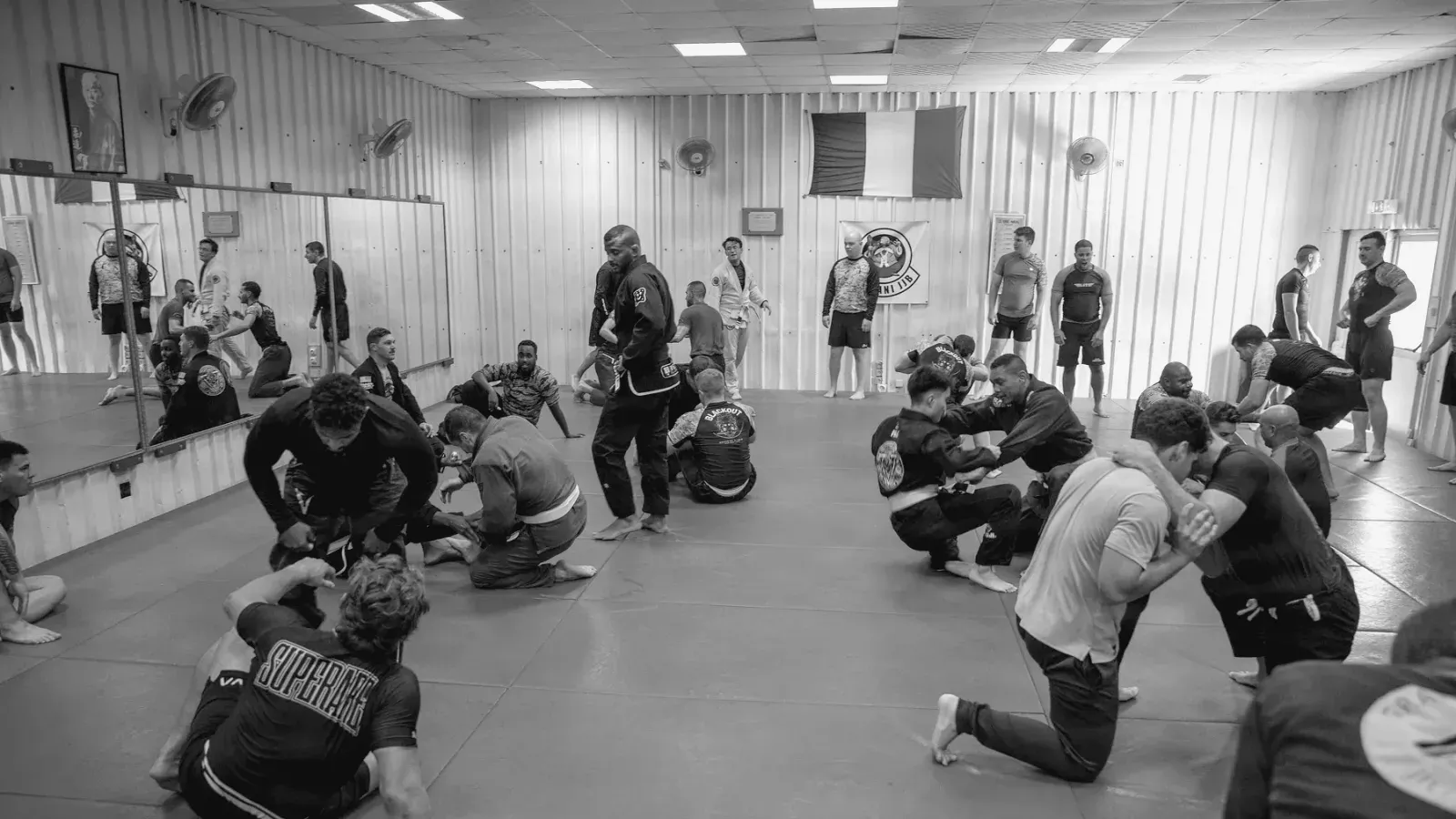 Wide view of a training hall with several pairs of students grappling on blue mats.
