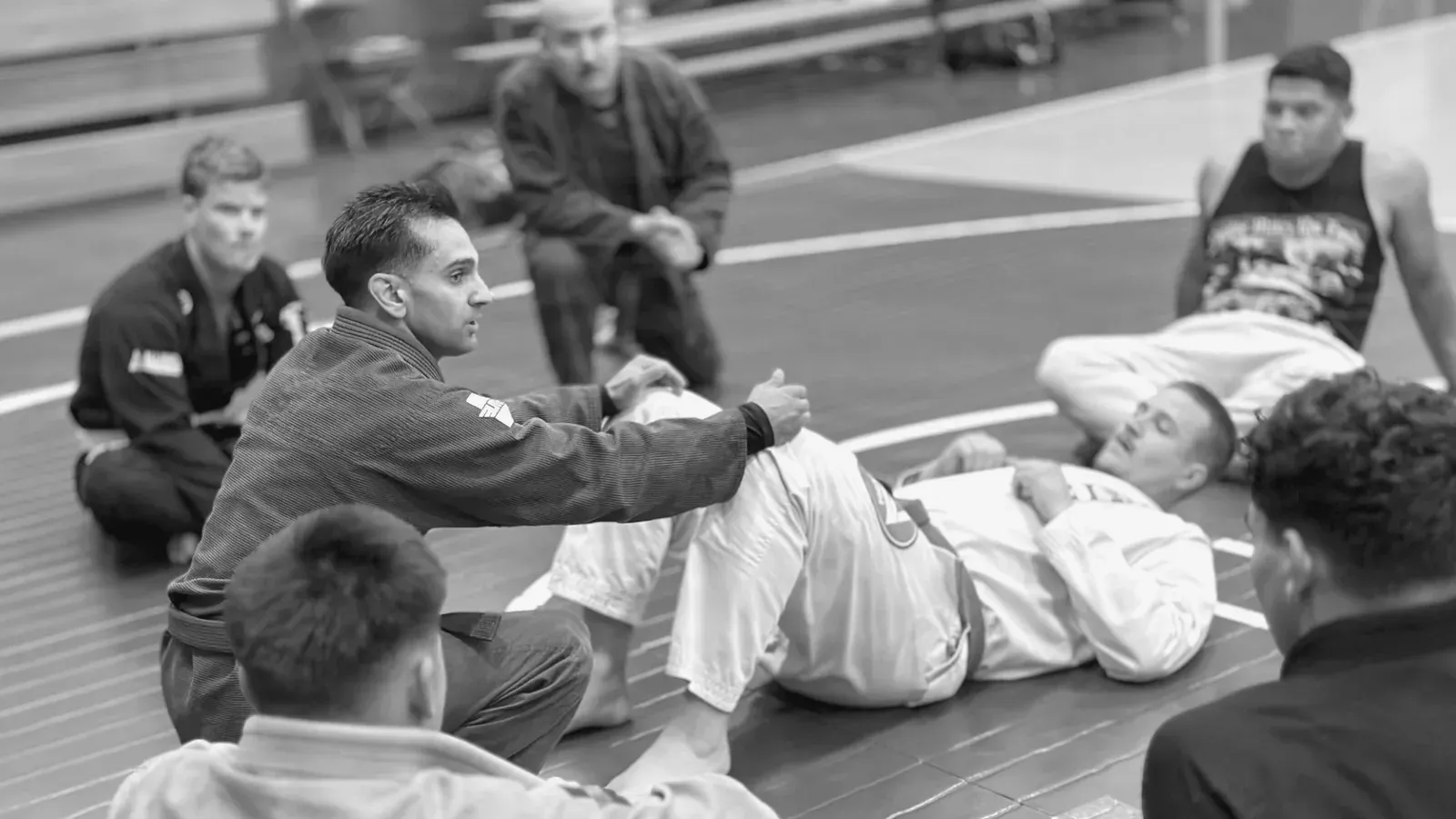A Brazilian Jiu Jitsu instructor demonstrating a ground position while a beginners’ class watches from around the mat.