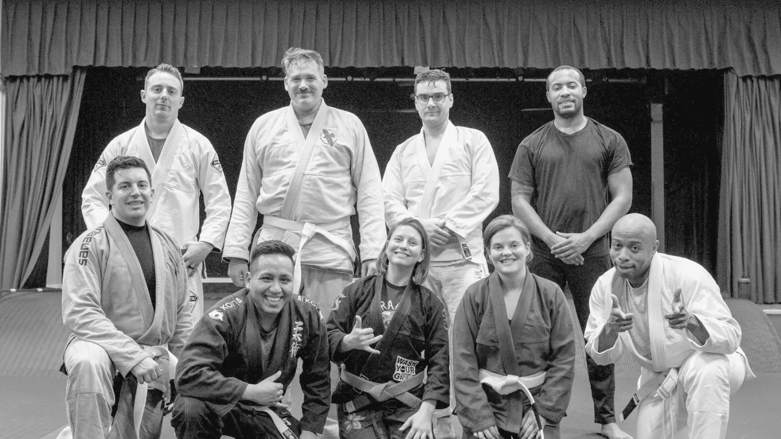 A group of Brazilian Jiu Jitsu students posed together on the mat in gis and rashguards, wearing belts of several different colours.