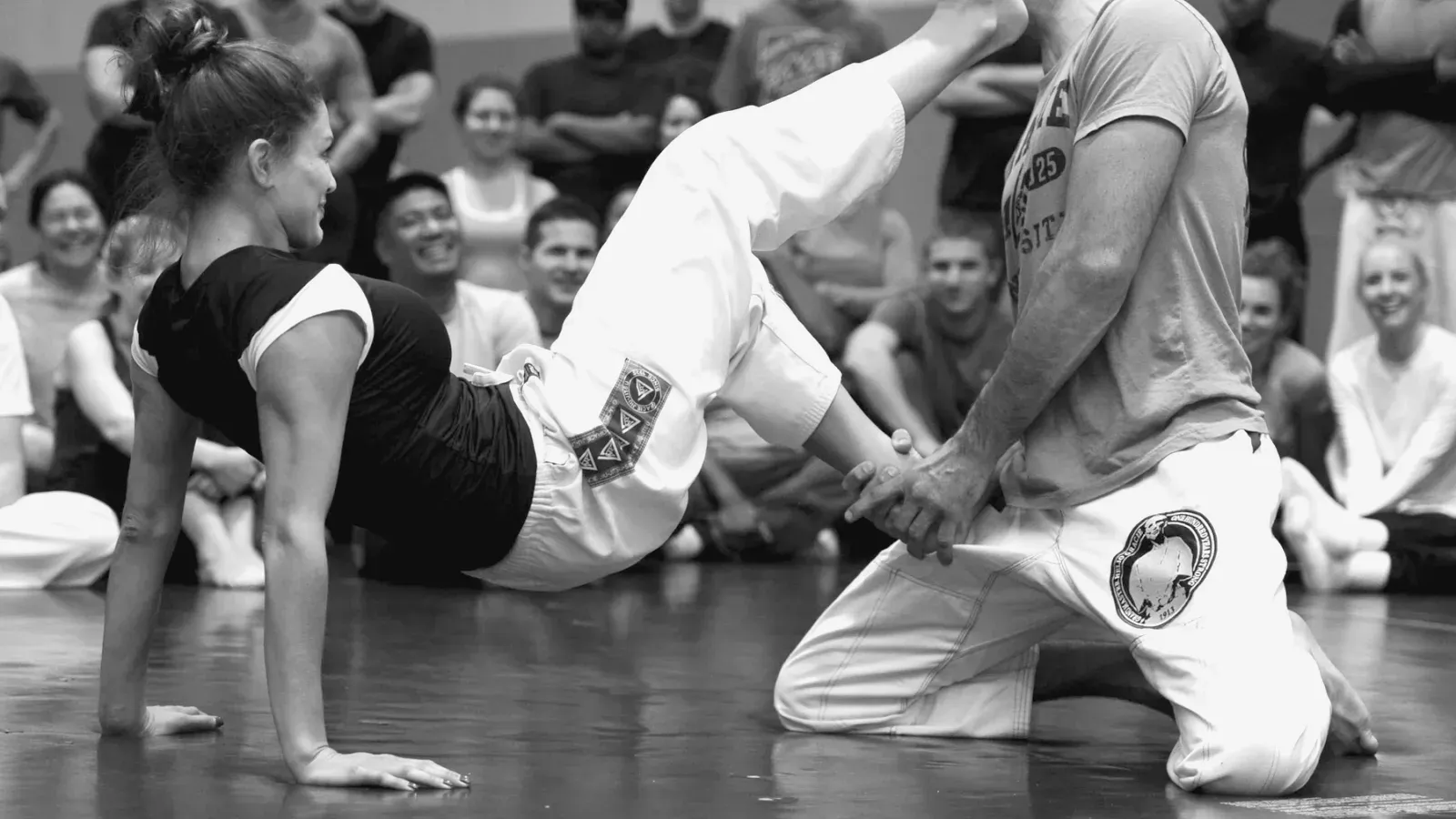 A Brazilian Jiu Jitsu instructor demonstrating on a partner while a class of students watches from the mat.