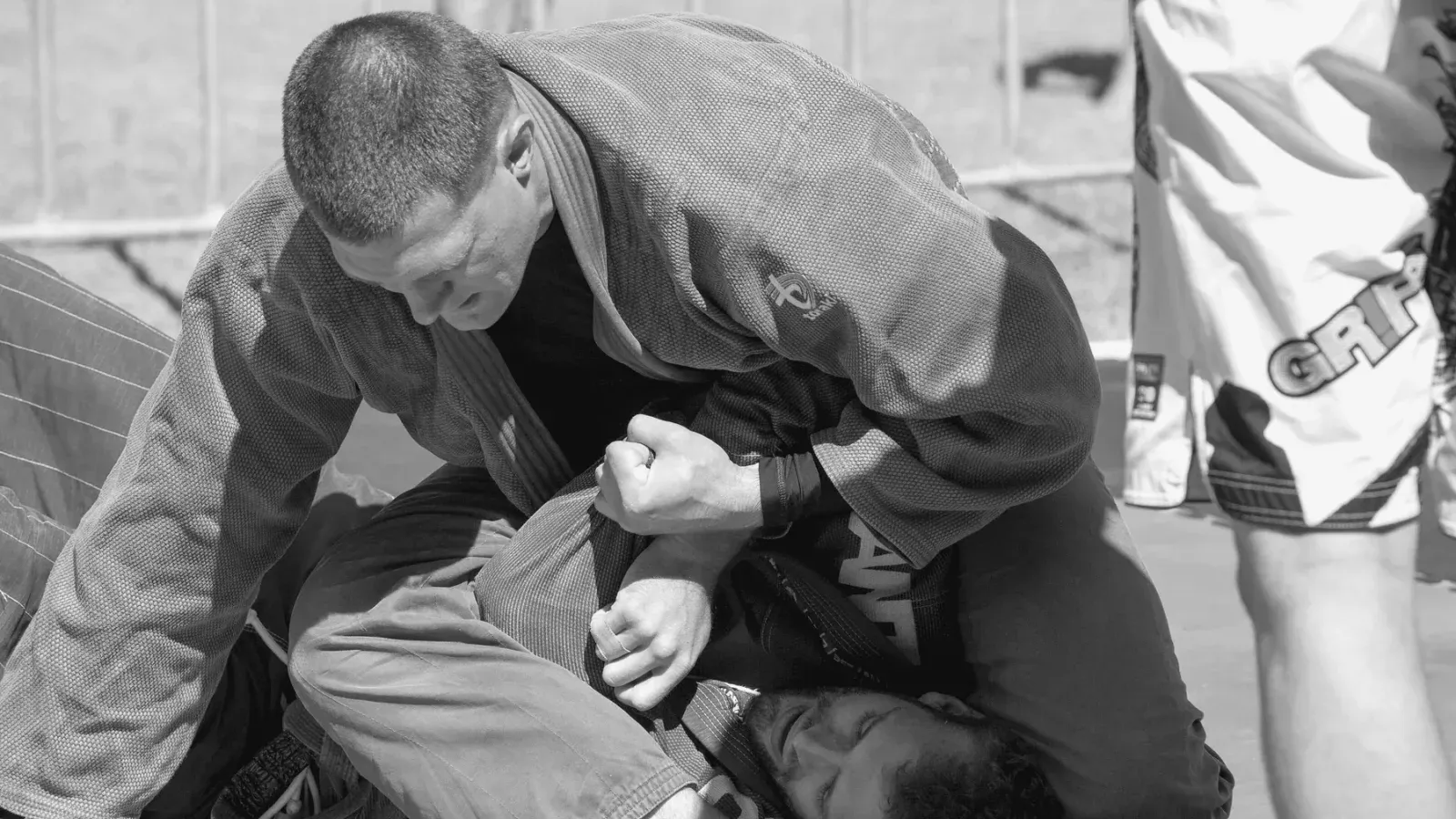 Two grapplers in gis working a ground position, one controlling the other from the top.
