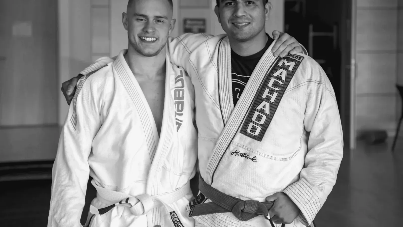 Two Brazilian Jiu Jitsu training partners standing side by side in gis at the academy, one wearing a white belt and one a purple belt.