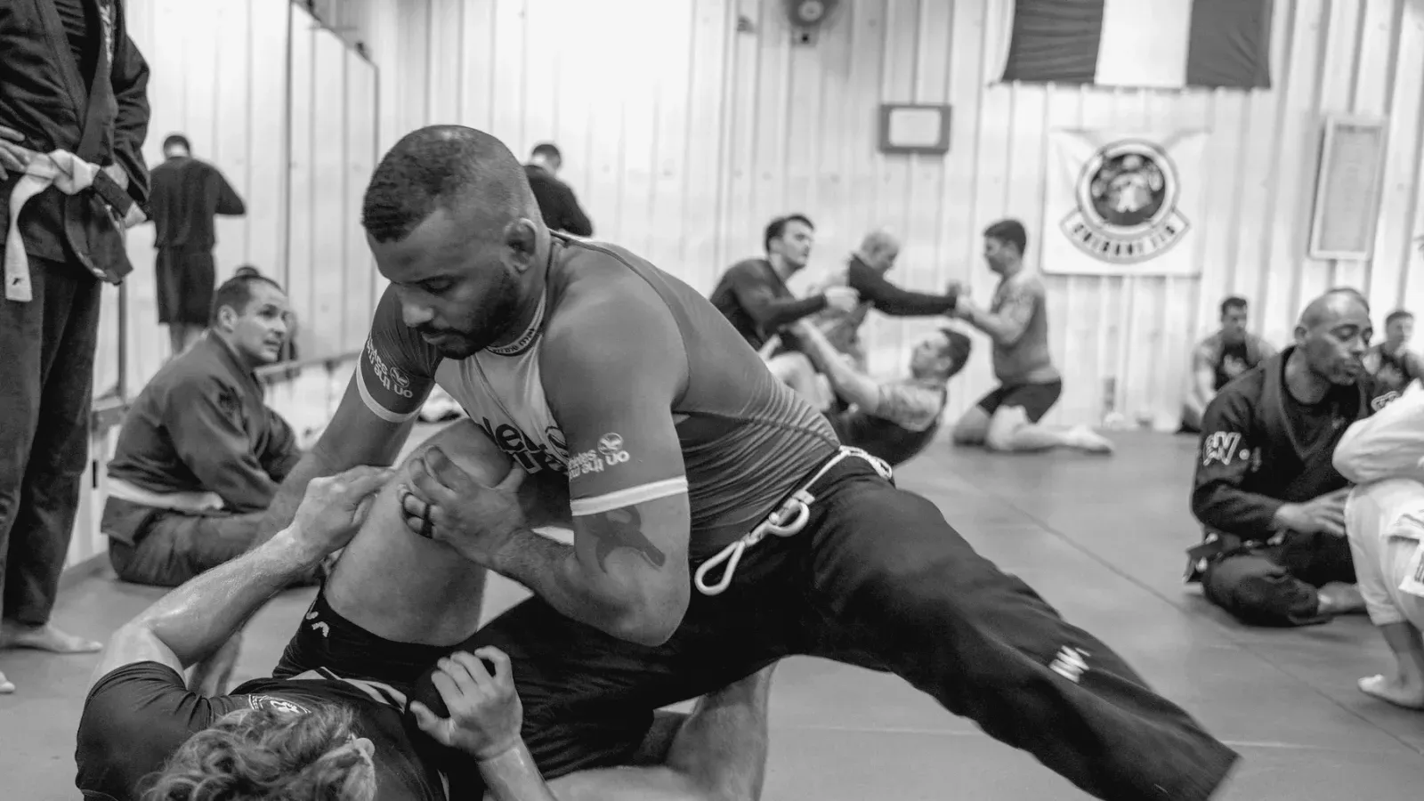 Two grapplers working from guard on the mat while other pairs train behind them in a busy academy.