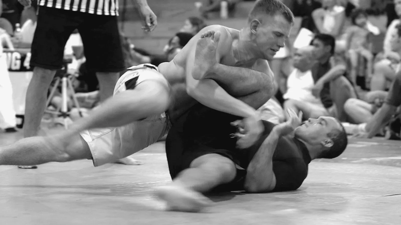 Two no-gi competitors on the mat, one playing guard from their back during a match.