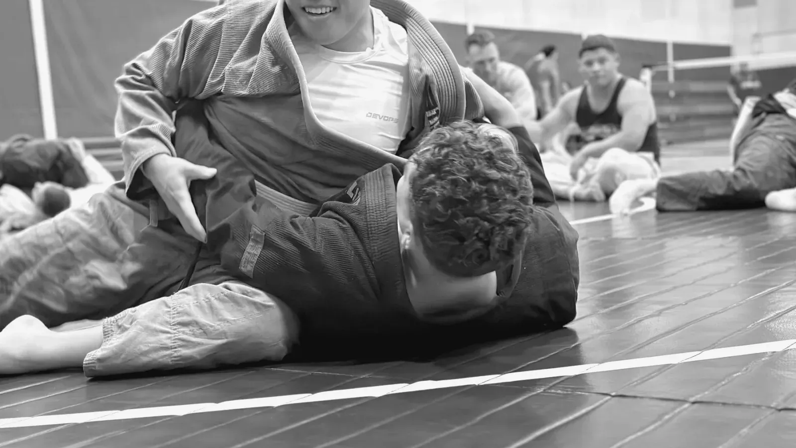 Two grapplers on a blue mat during a no-gi session, one holding a controlling position on the ground while others train behind them.