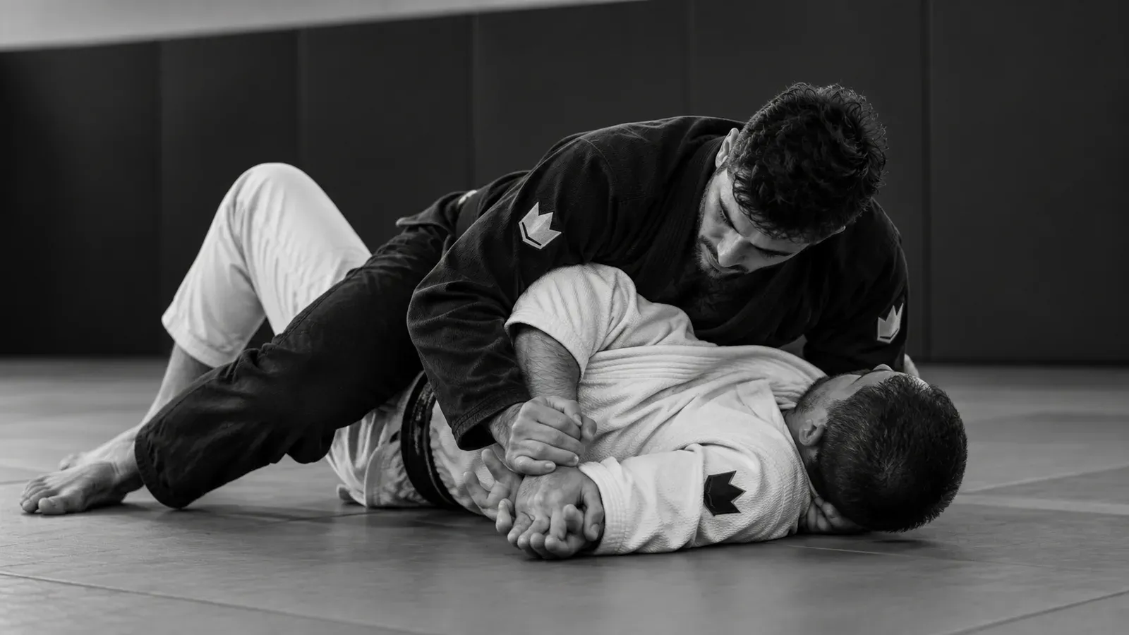 From side control, a grappler in a black gi controls the wrist of a partner in a white gi lying on the mat
