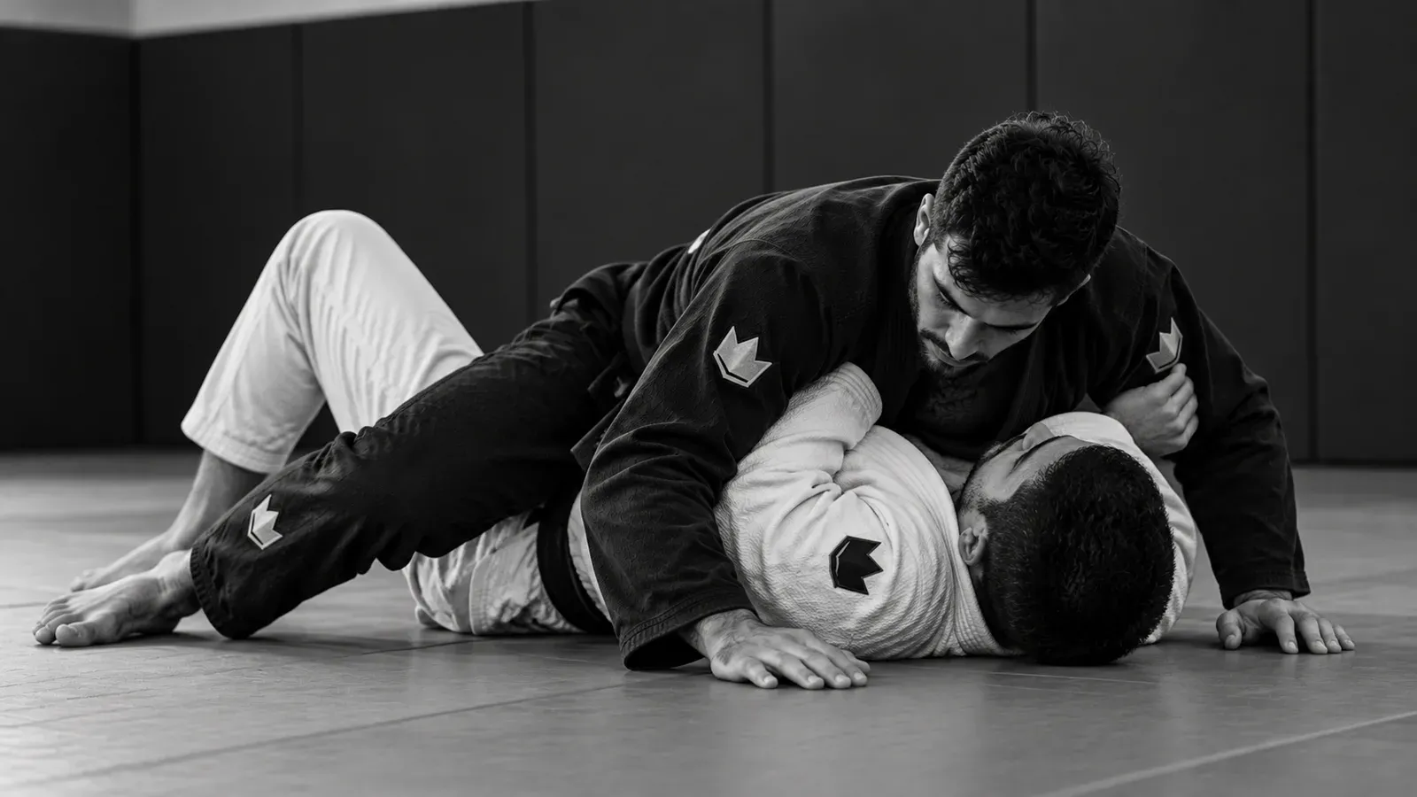 A grappler in a black gi pins a partner in a white gi chest to chest in side control on the mat