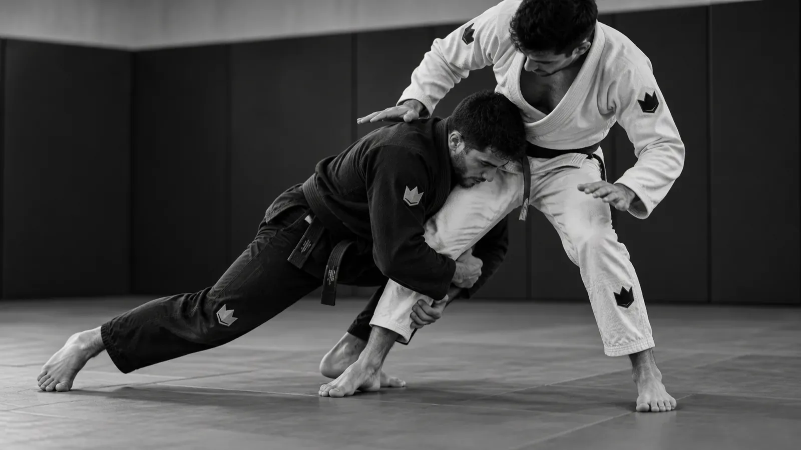 A grappler in a black gi drives in on the leg of a standing partner in a white gi for a takedown